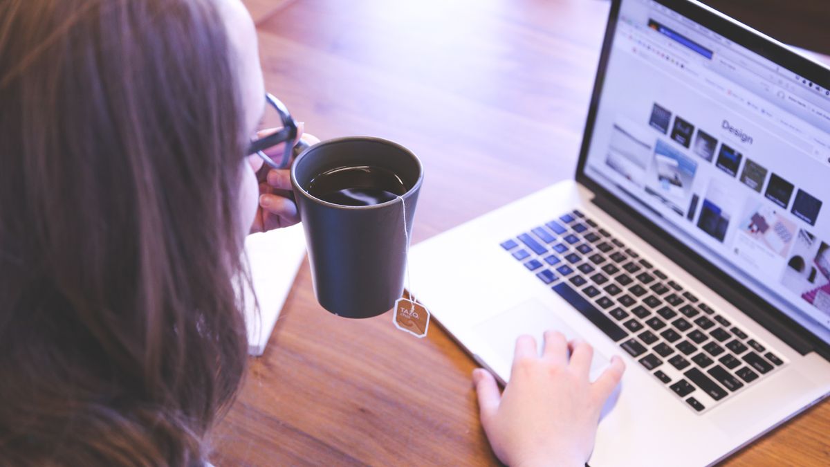 A woman enjoys coffee while browsing the web on a laptop at a home office desk