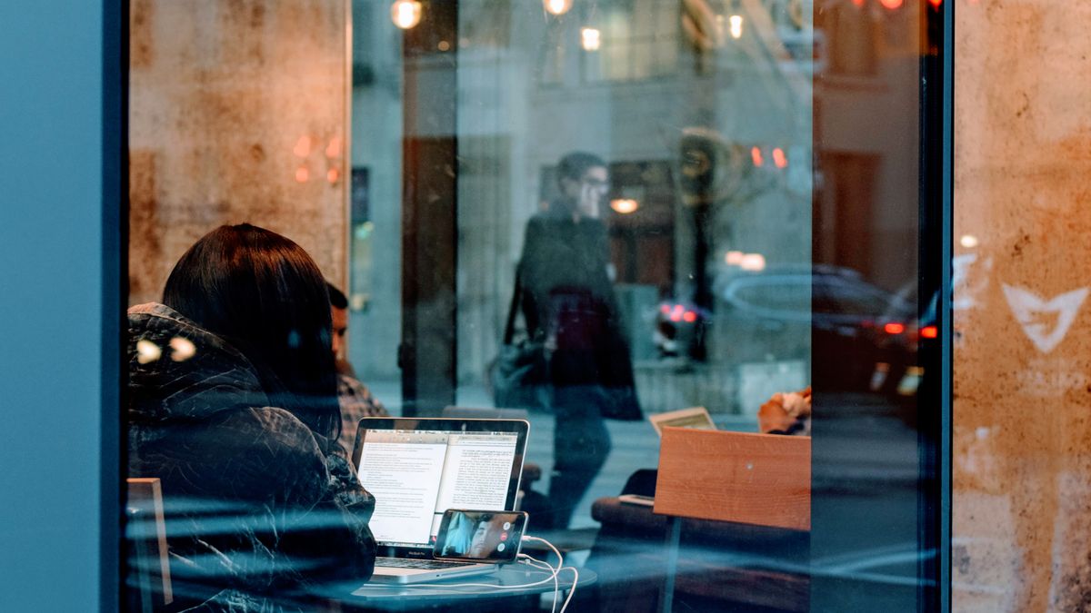A person working on a laptop inside a café, viewed through a window with street reflections