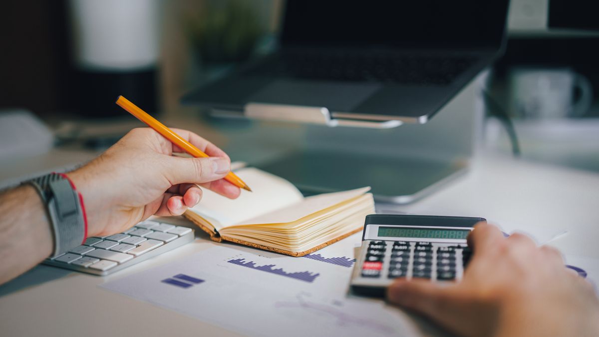 Hands using calculator and taking notes on desk with laptop, showcasing business analytics