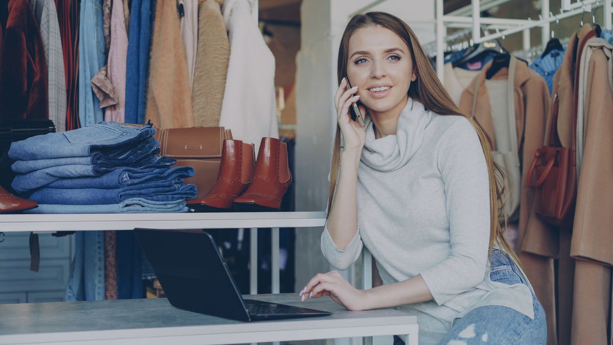 Woman in boutique talks on phone with laptop, surrounded by clothes